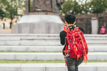 A young Asian male photographer with a bright backpack on his shoulders photographs the monument. Rear view