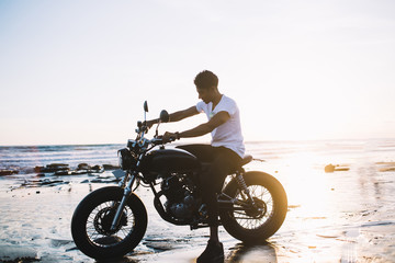 Fototapeta na wymiar Ethnic biker sitting on motorcycle on sea beach during sunset