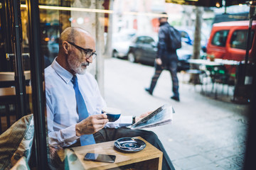 Senior focused businessman in glasses with coffee cup reading newspaper