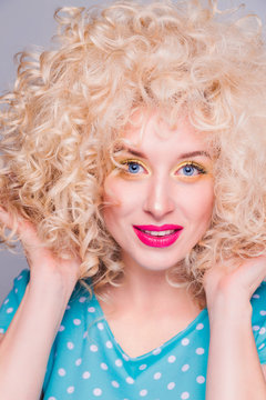 Beautiful blonde girl in retro style with voluminous curly hairstyle, in a blue polka-dot blouse on a gray background, enjoys her hairstyle and looks at the camera