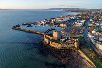 Medieval Norman Castle in Carrickfergus and Belfast Lough in sunrise light. Aerial view with marina, yachts, parking, breakwater, groyne, sediments and far view of Belfast in the background