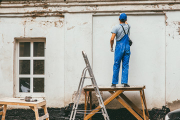 unrecognizable worker in blue construction pants stands on a wooden stand and works with a spatula with plaster on the wall. Renovation of the building's facade on the street