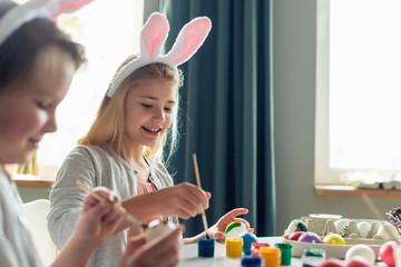 Girl wearing in bunny ears painting an Easter eggs and smiling. - Image