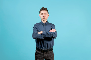 A teenager in a blue shirt with folded arms on his chest stands on a blue background with a serious face