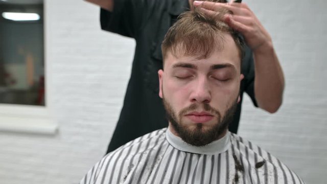 Man with his eyes closed sits in a barbershop chair and cuts his hair at a male hairdresser.Relaxed bearded client uses the services of a professional barber.Process creating a stylish men's hairstyle