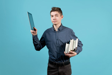 A teenager in a blue shirt holds a tablet and a stack of books under his armpit. Raises the tablet...