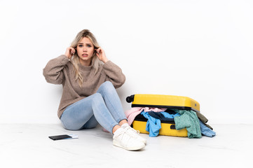 Teenager blonde girl with a suitcase full of clothes sitting on the floor frustrated and covering...