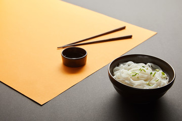 selective focus of rice noodles in bowl near chopsticks and soy sauce on yellow and black surface