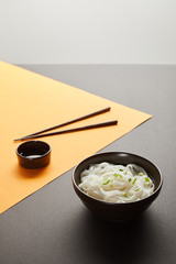 selective focus of rice noodles in bowl near chopsticks and soy sauce on yellow and black surface isolated on grey