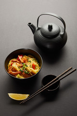 noodles with shrimps and vegetables in bowl near lemon, teapot, wooden chopsticks and soy sauce on black background