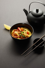 noodles with shrimps and vegetables in bowl near lemon, teapot, wooden chopsticks and soy sauce on black background