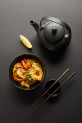 top view of noodles with shrimps and vegetables in bowl near lemon, teapot, wooden chopsticks and soy sauce on black background