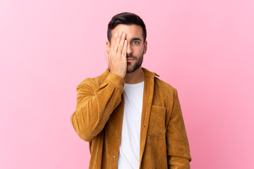 Young handsome man with beard wearing a corduroy jacket over pink background covering a eye by hand