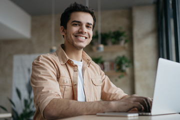Portrait of happy Indian student studying, learning language. Mixed race business man using laptop computer, typing on keyboard, looking at camera and smiling. Copywriter working freelance project - obrazy, fototapety, plakaty