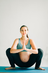 a young pretty pregnant girl in sports clothes is doing yoga, doing asana Malasana on the Mat in the Studio