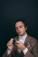 A dapper young man obviously you wearing a white turtleneck sweater and a beige jacket with handkerchief in his top breast pocket, looking pensively at his wrist watch, contemplating time