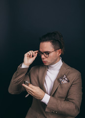 A dapper young man obviously you wearing a white turtleneck sweater and a beige jacket with handkerchief in his top breast pocket, looking pensively at his wrist watch, contemplating time