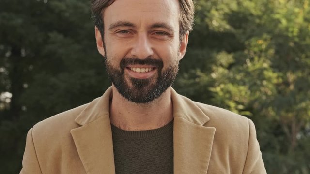 Happy elegant bearded man in coat looking at the camera outdoors
