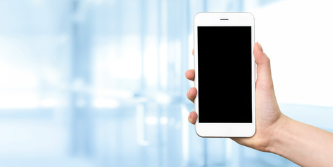 Woman hand holding a white smartphone with blank screen on a blurred background of a modern corporate interior with copy space