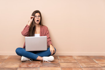 Teenager student girl sitting on the floor with a laptop with glasses and surprised