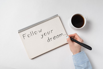 Sheet of paper with the words Follow your dream. Female hand writing in notebook, cup of coffee. Top view white background
