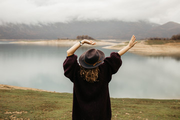 Happy beautiful woman in purple sweater holding her hat and enjoying trip on the lake. Female tourist exploring lake. She is standing turned back and looking into distance, with her hands in the air.
