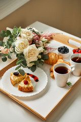 Fototapeta na wymiar Wholesome breakfast on a wooden tray. Croissants and cheesecakes with tea and fresh berries are served on a tray in bed for a loved one.