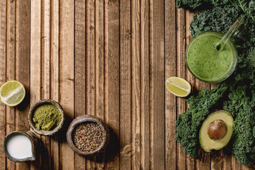 Glass of green healthy vegan smoothie, glass straw. Ingredients above. Kale, bananas, avocado, lime, non-diary milk, matcha powder and seeds over wooden plank background. Flat lay, space