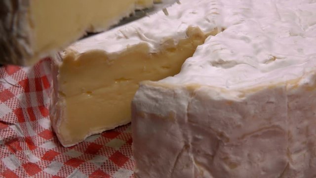 Piece of soft camambert cheese is taken out with a knife from a cheese wheel on a red-white vichy napkin