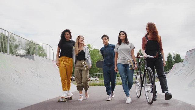Female Friends With Skateboards And Bike Walking Through Urban Skate Park