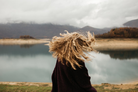 happy girl with curly blond hair dances on a lake alone, her hair is flying because of the wind flow, free as a bird. photo of girl with curly hair standing back to camera over lake background.