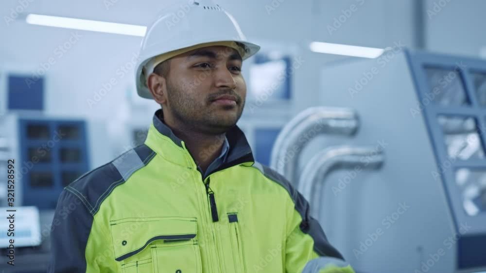 Poster Industry 4 Factory: Portrait of a Modern Worker Wearing Safety Jacket Puts on Hard Hat and Walks Through Contemporary Industrial Workshop where Professionals are Working and Programming CNC Machinery