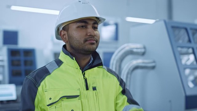 Industry 4 Factory: Portrait of a Modern Worker Wearing Safety Jacket Puts on Hard Hat and Walks Through Contemporary Industrial Workshop where Professionals are Working and Programming CNC Machinery