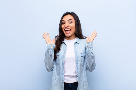 young latin pretty woman looking happy and excited, shocked with an unexpected surprise with both hands open next to face against flat wall