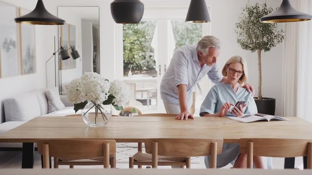 Senior Couple In Dining Room At Home Looking At And Mobile Phone Together