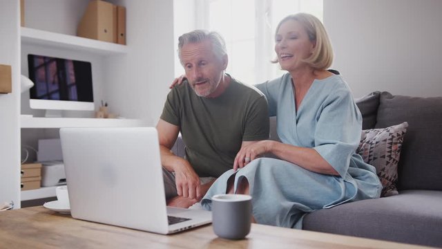 Senior Couple Sitting On Sofa At Home Making Video Call Using Laptop Computer Together