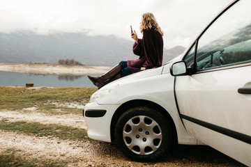 Beautiful woman with curly hair, sitting on the car, holding mobile phone and taking photos of lake on a cloudy day. She is texting on smartphone and taking selfie. 