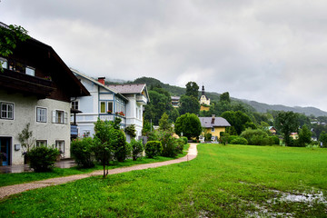 Fototapeta na wymiar Mondsee, Austria - A row of houses in the countryside, next to a green lawn, in the background you can see a church standing on a hill among green trees, in the summer afternoon.