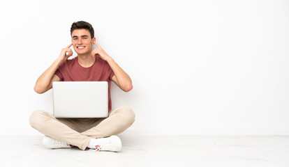 Teenager man sitting on the flor with his laptop frustrated and covering ears