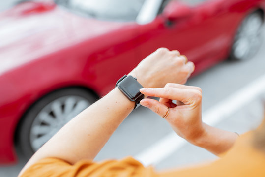 Woman with smartwatch at the car parking with red vehicle on the background, close-up view on watch with black screen. Car control with a smart watch concept