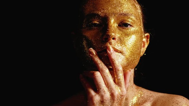 Glitter art portrait. Organic face mask. Woman touching golden sparkling skin over black background.