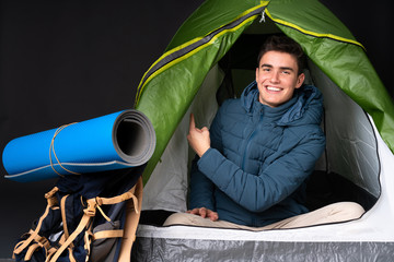 Teenager caucasian man inside a camping green tent isolated on black background pointing back