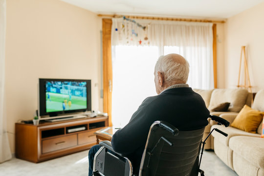 Back View Of Senior Man Sitting In Wheelchair Watching TV At Home