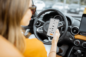 Fototapeta na wymiar Young woman controlling car using smart phone with launched mobile application, sitting on the driver's seat, view from the backside