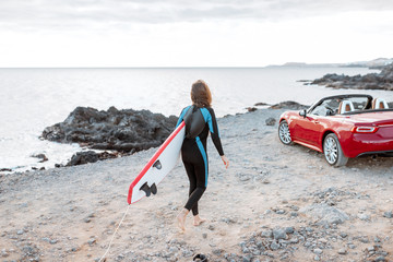 Young woman surfer walking with surfboard on the rocky ocean coast near her sports car. Active...