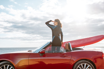 Woman enjoying beautiful landscapes while standing on her sports car with a surfboard near the ocean. Carefree lifestyle and active sports concept