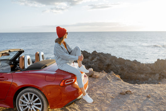 Woman enjoying beautiful view on the ocean, sitting on the convertible car on the rocky coast during a sunset. Carefree lifestyle and car travel concept