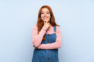 Teenager redhead girl with overalls over isolated blue background laughing