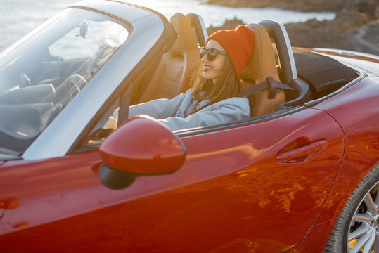 Lifestyle portrait of happy woman in red hat driving convertible car on the rocky ocean coast during a sunset. Carefree lifestyle and car travel concept © rh2010