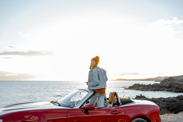 Woman enjoying beautiful view on the ocean, sitting on the convertible car on the rocky coast during a sunset. Wide view with space on the sky